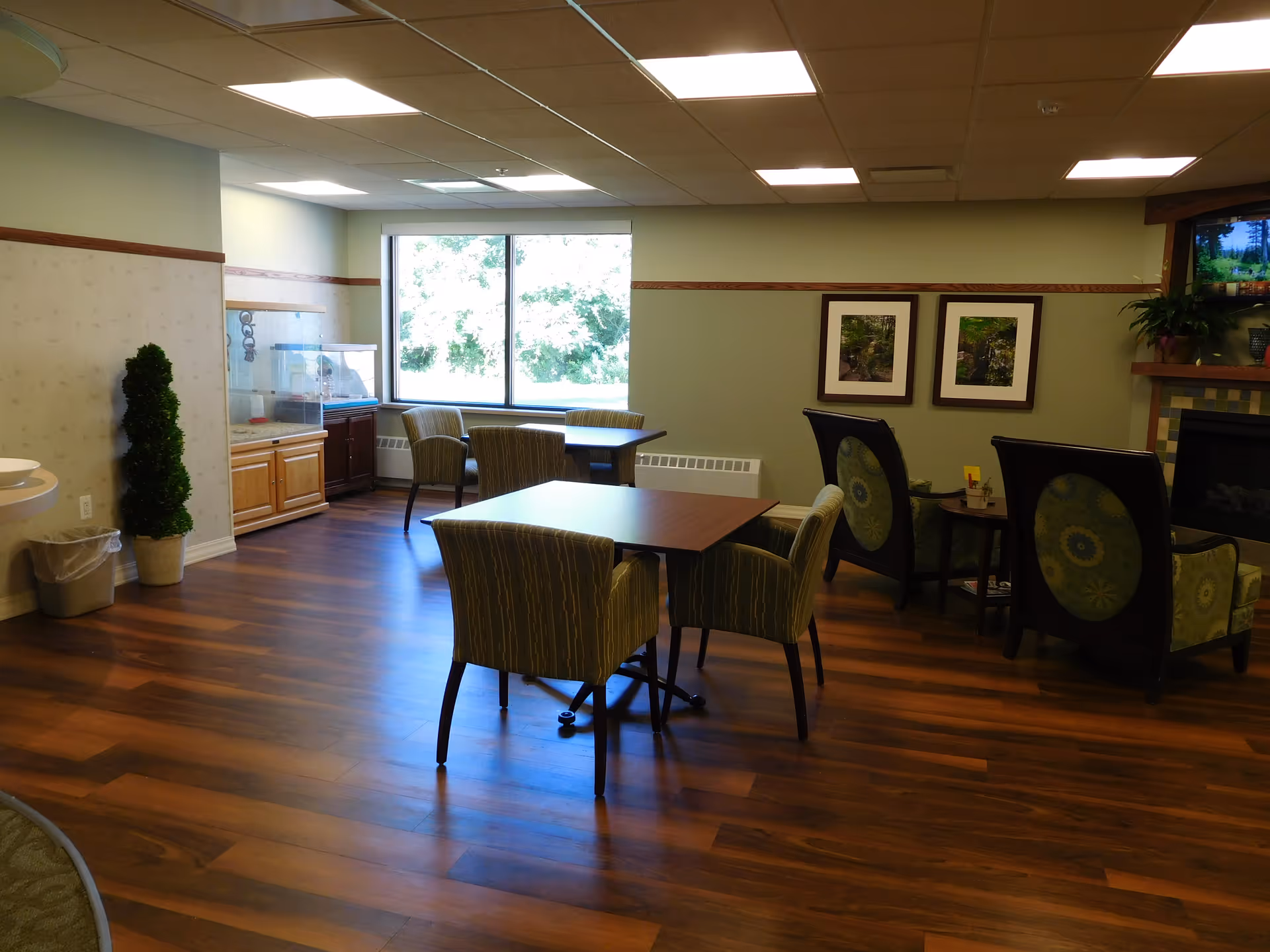 A cozy common area in a senior living facility with wooden flooring, several tables and chairs, a large window letting in natural light, framed pictures on the wall, a fireplace with a TV above it, and a small potted plant near a trash can.