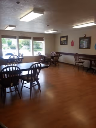 Interior view of a senior care facility dining room with wooden tables and chairs arranged neatly. Large windows allow natural light to brighten the room, and framed pictures hang on the walls.