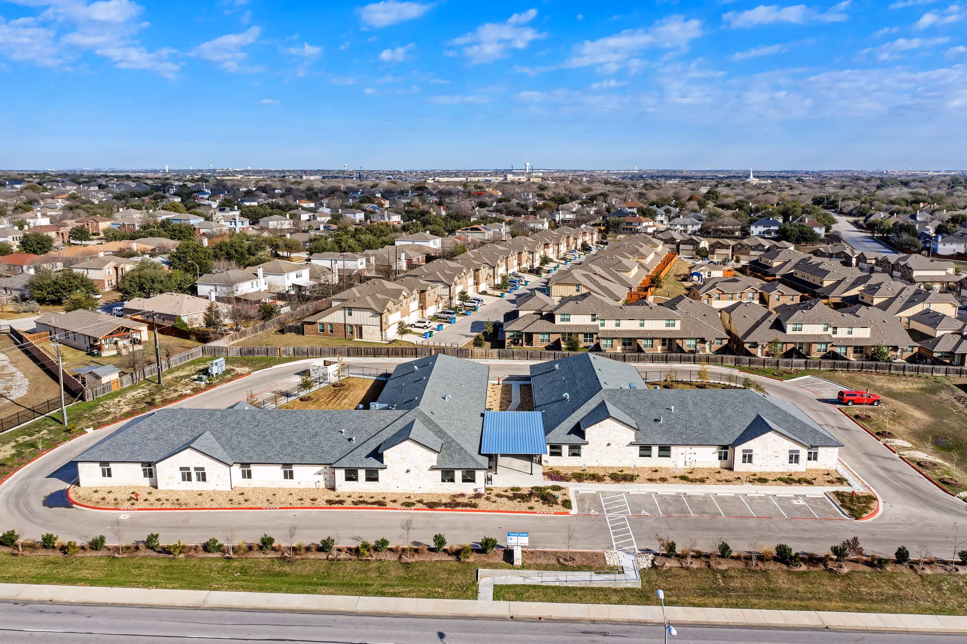 Aerial view of a single-story assisted living facility with parking, landscaping, and a surrounding residential neighborhood under a blue sky.