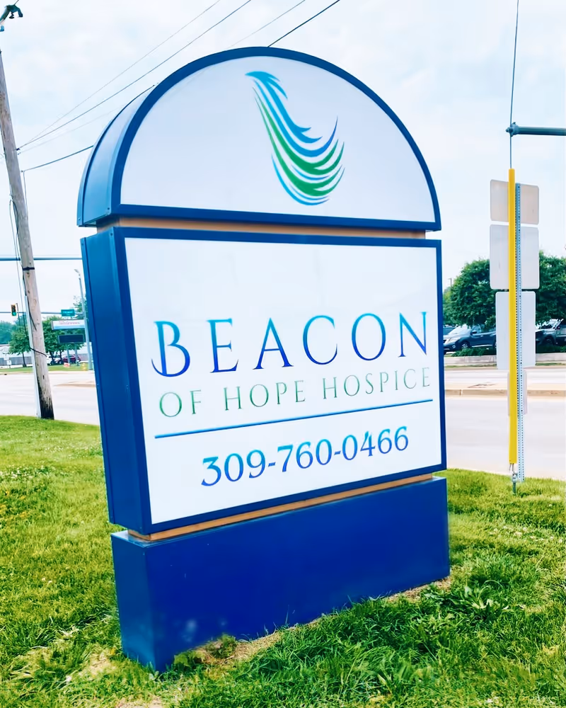 Large blue beacon sign for Beacon of Hope Hospice of Illinois on a grassy roadside.
