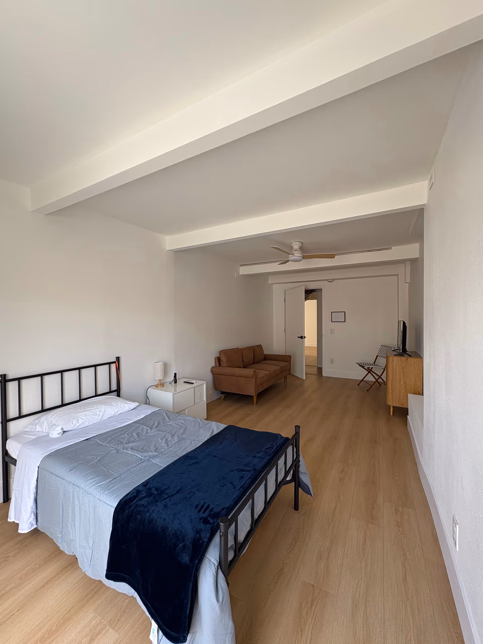 A spacious bedroom with metal bed, nightstand, couch, TV, and wooden floor.