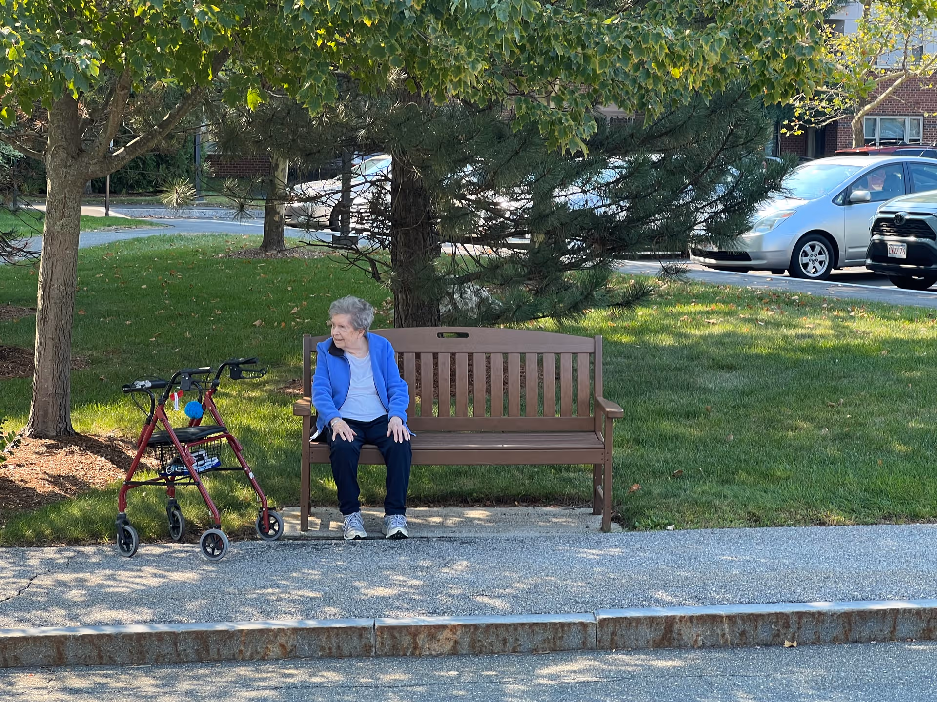Senior woman sits on a wooden park bench under trees with a walker nearby.
