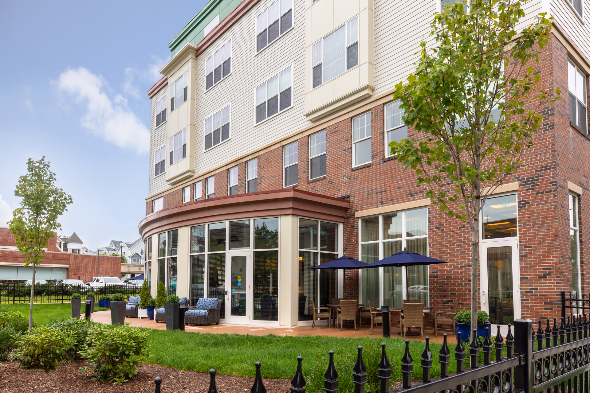 Exterior view of a multi-story senior living facility building with a brick and beige facade. The image shows a patio area with outdoor seating, including chairs and tables with blue umbrellas, surrounded by green grass, small trees, and shrubs. A black metal fence borders the garden area.