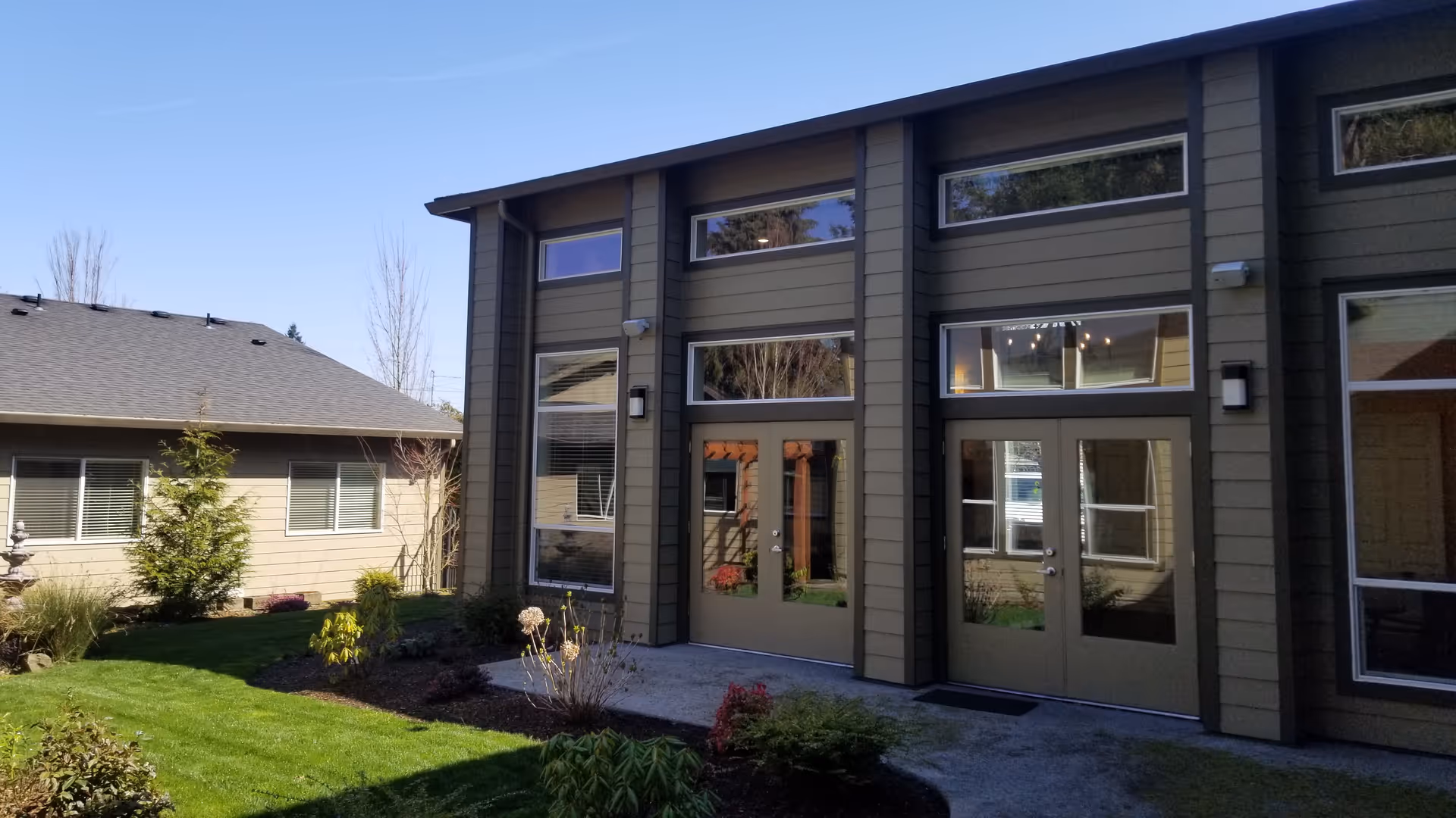 Courtyard view of a two-story senior living building with green siding, large windows and glass doors opening onto a landscaped lawn.