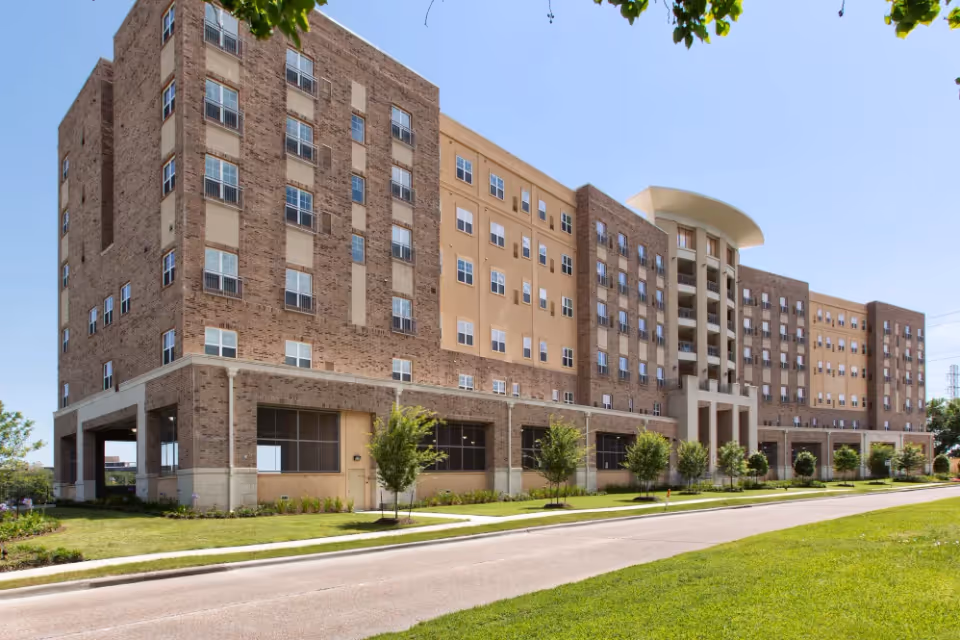 Multi-story brick and stucco senior living building with a curved entrance, rows of windows, and a landscaped lawn and driveway.