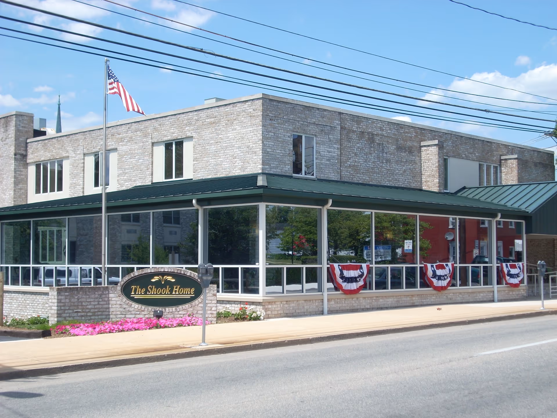 Exterior view of a two-story brick senior living facility with a glass-enclosed porch, American flag, and a sign reading "The Shook Home".