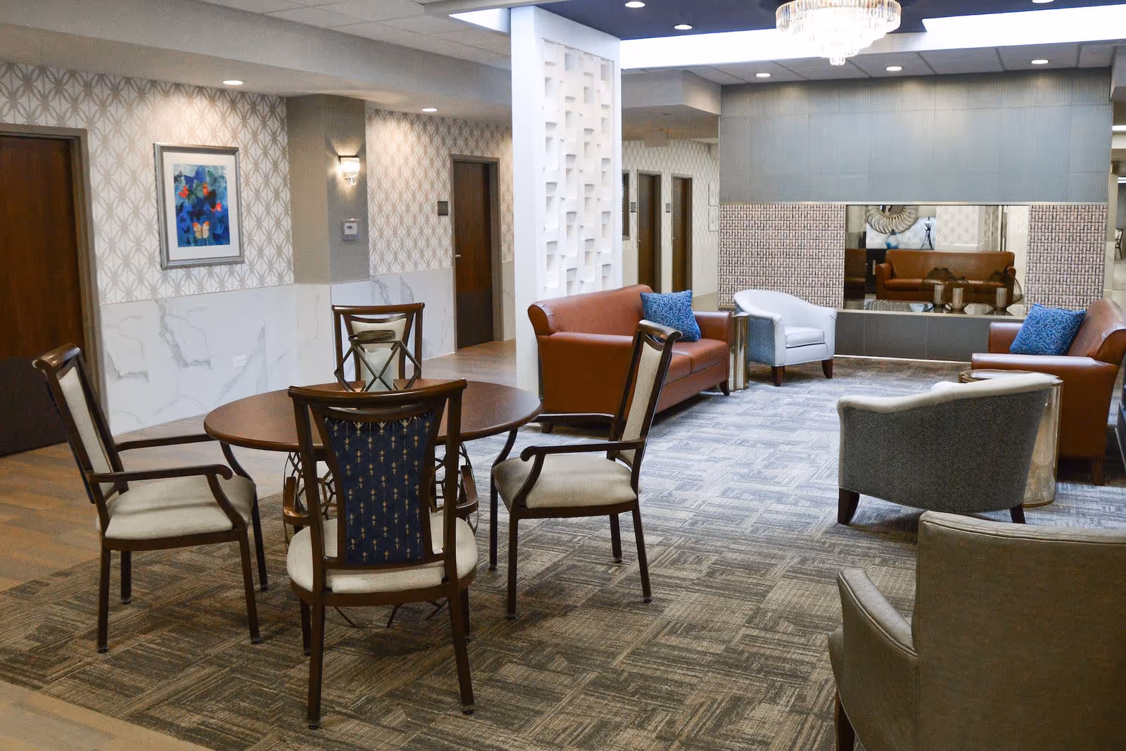 A spacious and well-lit common area in a supportive living facility featuring a round wooden table with four chairs, two brown leather sofas with blue accent pillows, two gray armchairs, a large decorative mirror on the wall, patterned wallpaper, and a chandelier overhead.