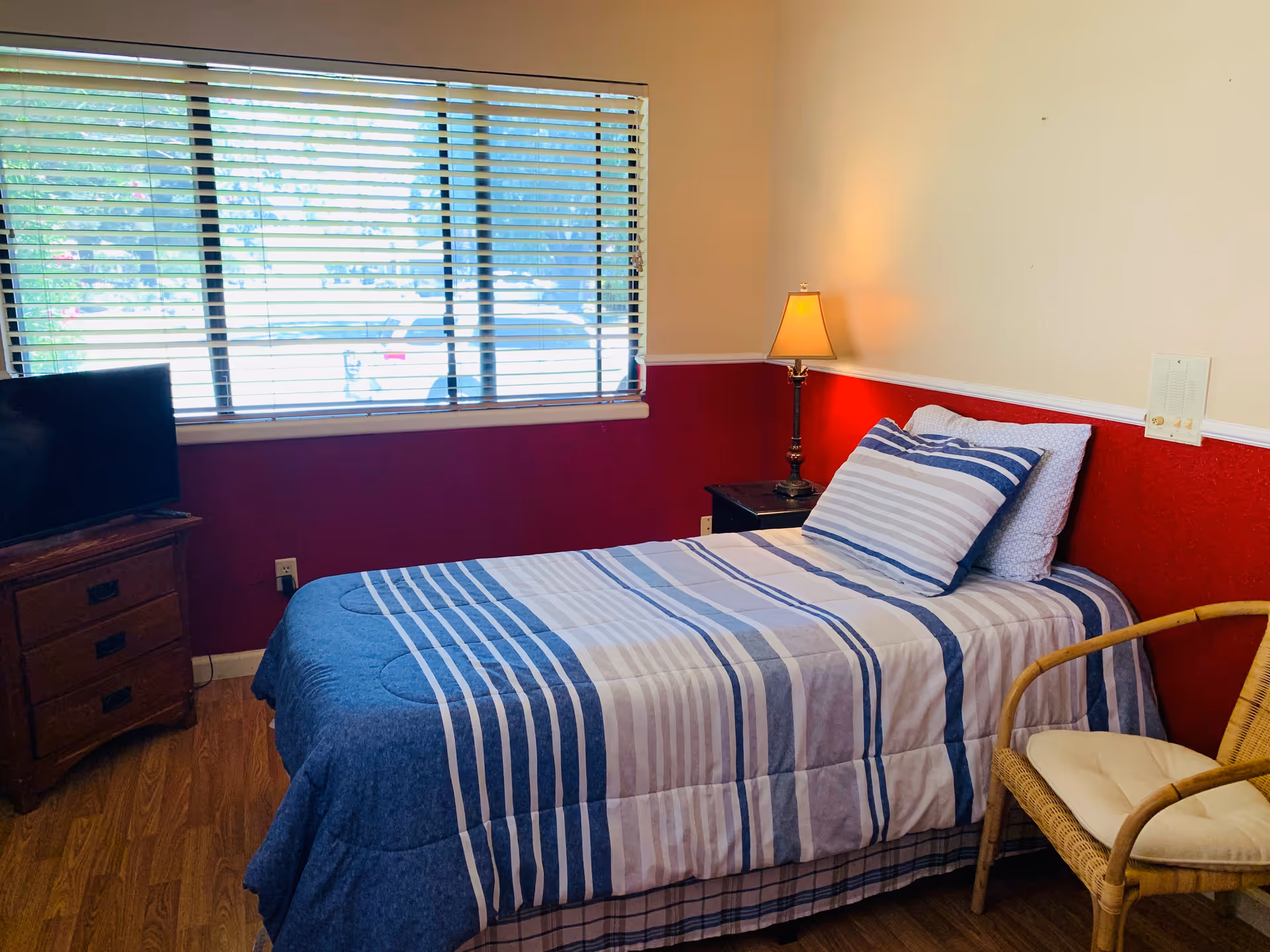 A tidy bedroom with a striped blue bedspread, window blinds, a nightstand lamp, and wicker chair.