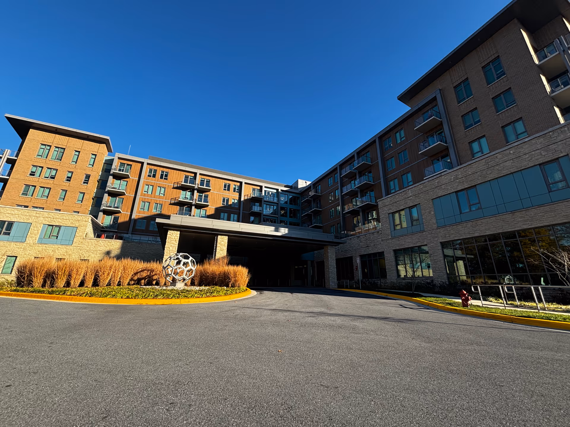 Exterior view of The Seneca Rockville building under a clear blue sky, showing a multi-story structure with balconies, large windows, and a covered entrance with a circular driveway. There is a modern metal sculpture surrounded by ornamental grasses near the entrance.