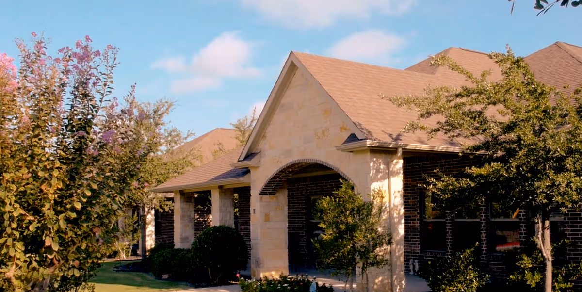 Single-story assisted living building with a stone archway entrance, brick facade, surrounding trees, and a blue sky.