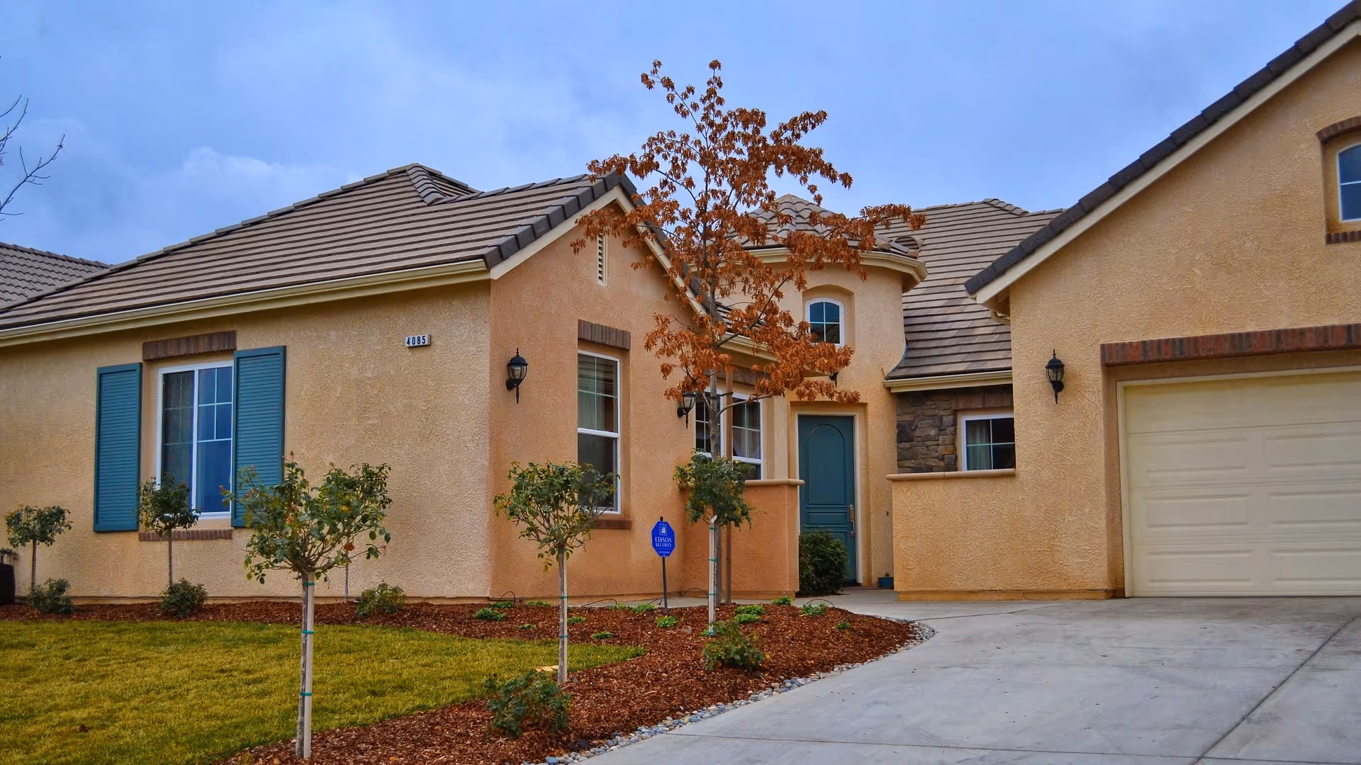 Front view of a single-story stucco house with a garage, driveway, small landscaped yard and a tree.