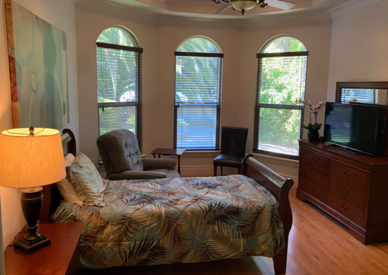 Bedroom in a senior living facility with a bed, dresser, TV, and large arched windows.