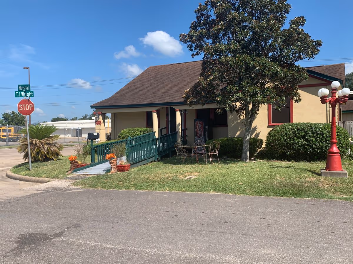 Single-story beige building with a green wheelchair ramp, front porch seating, a tree and a stop sign at the corner lot.