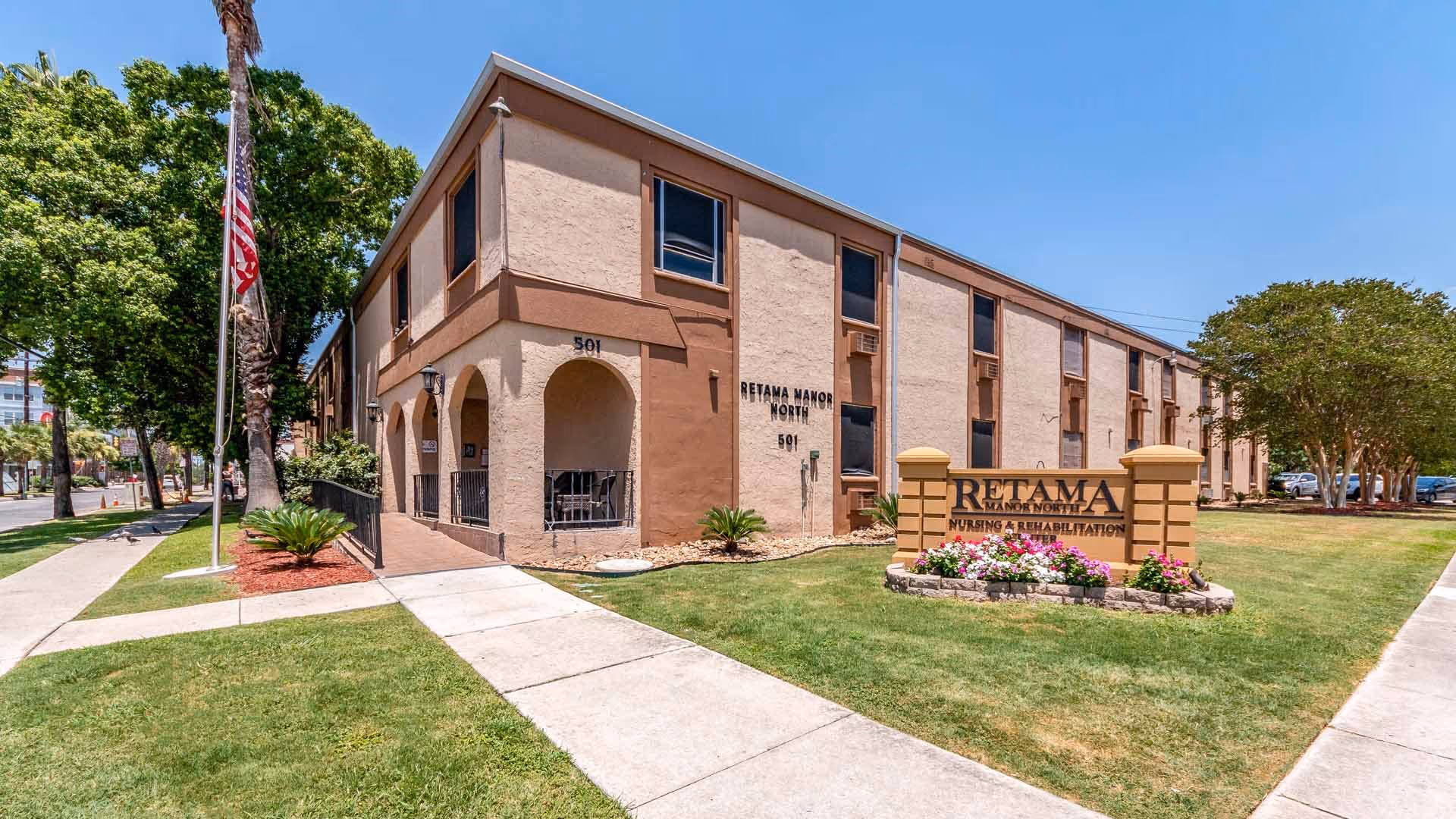 Two-story beige nursing and rehabilitation building with a front lawn, sidewalk, American flag, and a sign reading "RETAMA".