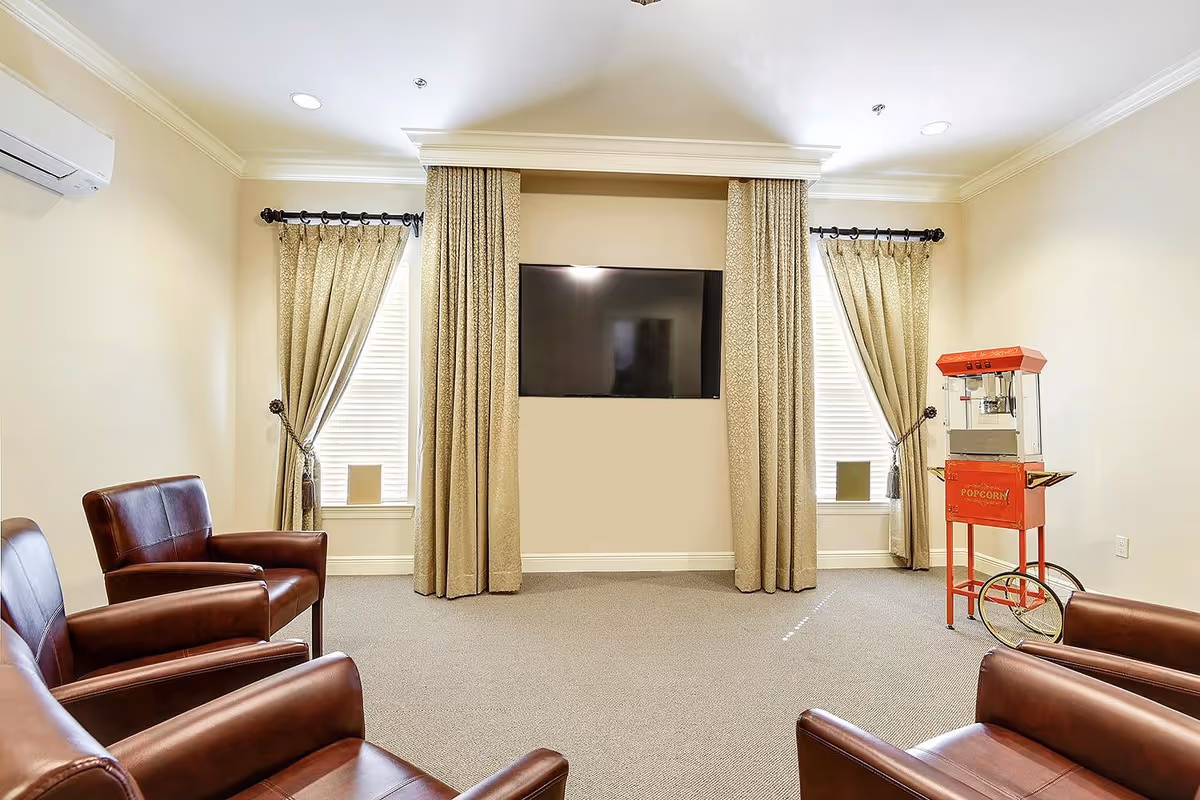 A cozy senior living common room with four brown leather armchairs arranged facing a wall-mounted flat screen TV. The room has beige walls, two windows with beige patterned curtains, and a red vintage-style popcorn machine on wheels in the corner.