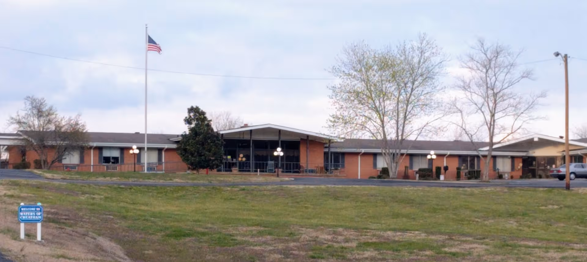 Single-story brick senior living facility with a front entrance, flagpole, and surrounding lawn.