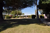 Front lawn and entrance of a single-story assisted living building flanked by trees and hedges under a clear sky.