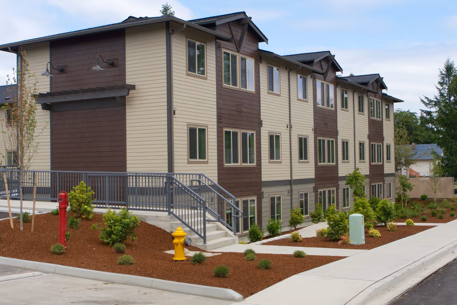 Three-story beige-and-brown apartment building with multiple windows, landscaped beds, a sidewalk, and visible red and yellow fire hydrants.