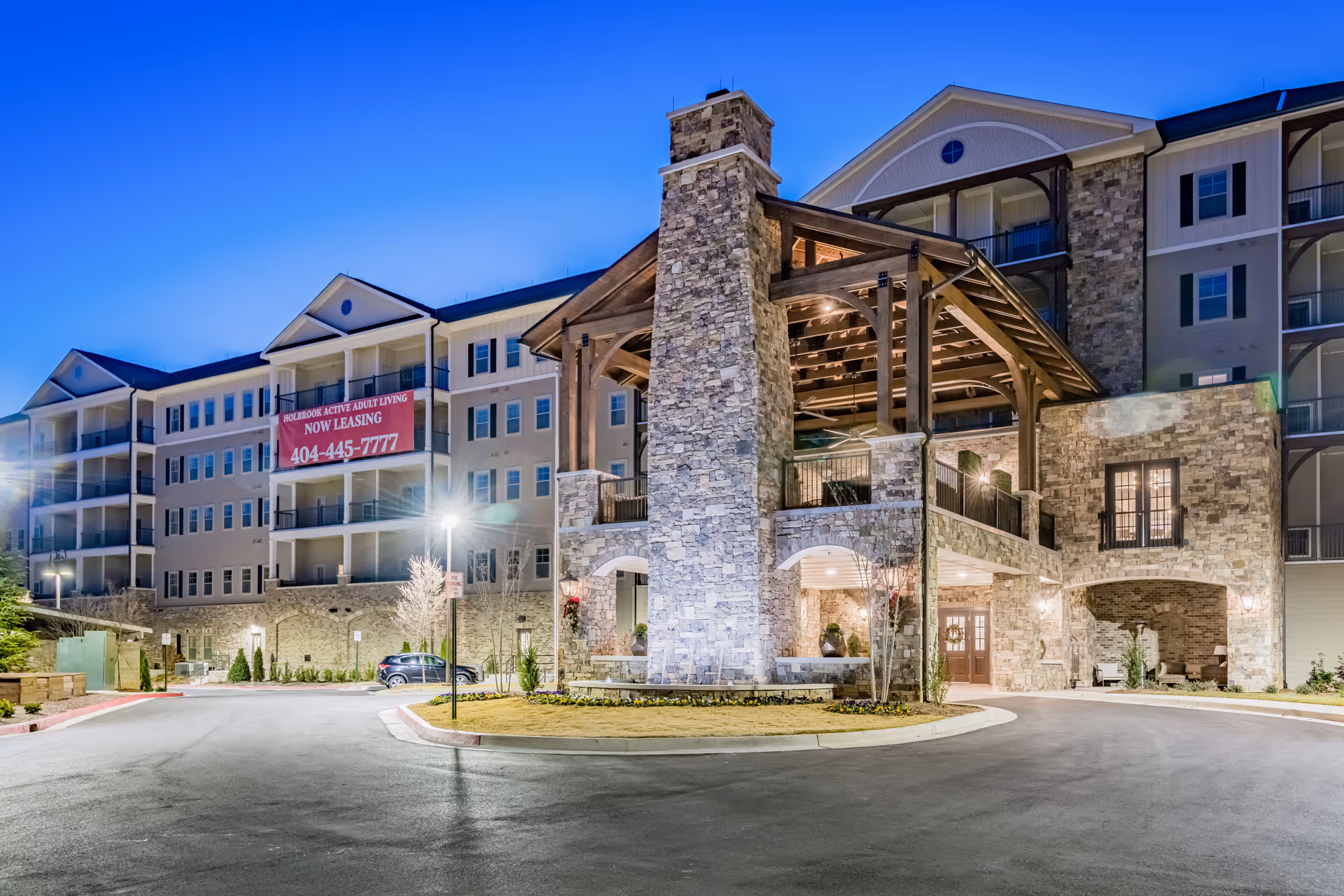 Front exterior of a multi-story stone-clad senior living building with a covered entrance illuminated at dusk.