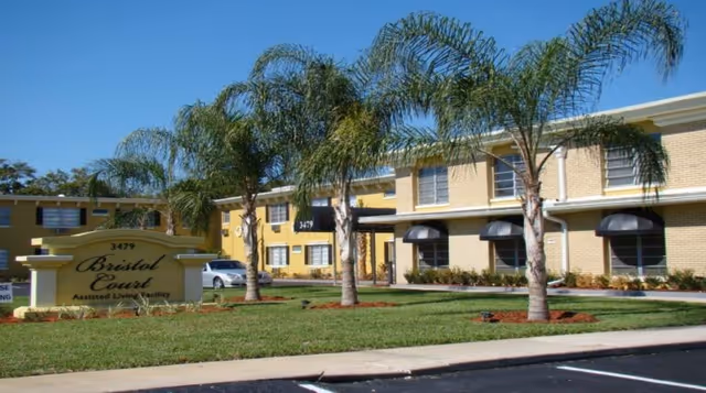 Exterior view of a two-story assisted living facility named Bristol Court with yellow brick walls, several palm trees in front, and a sign displaying the facility name and address 3479.