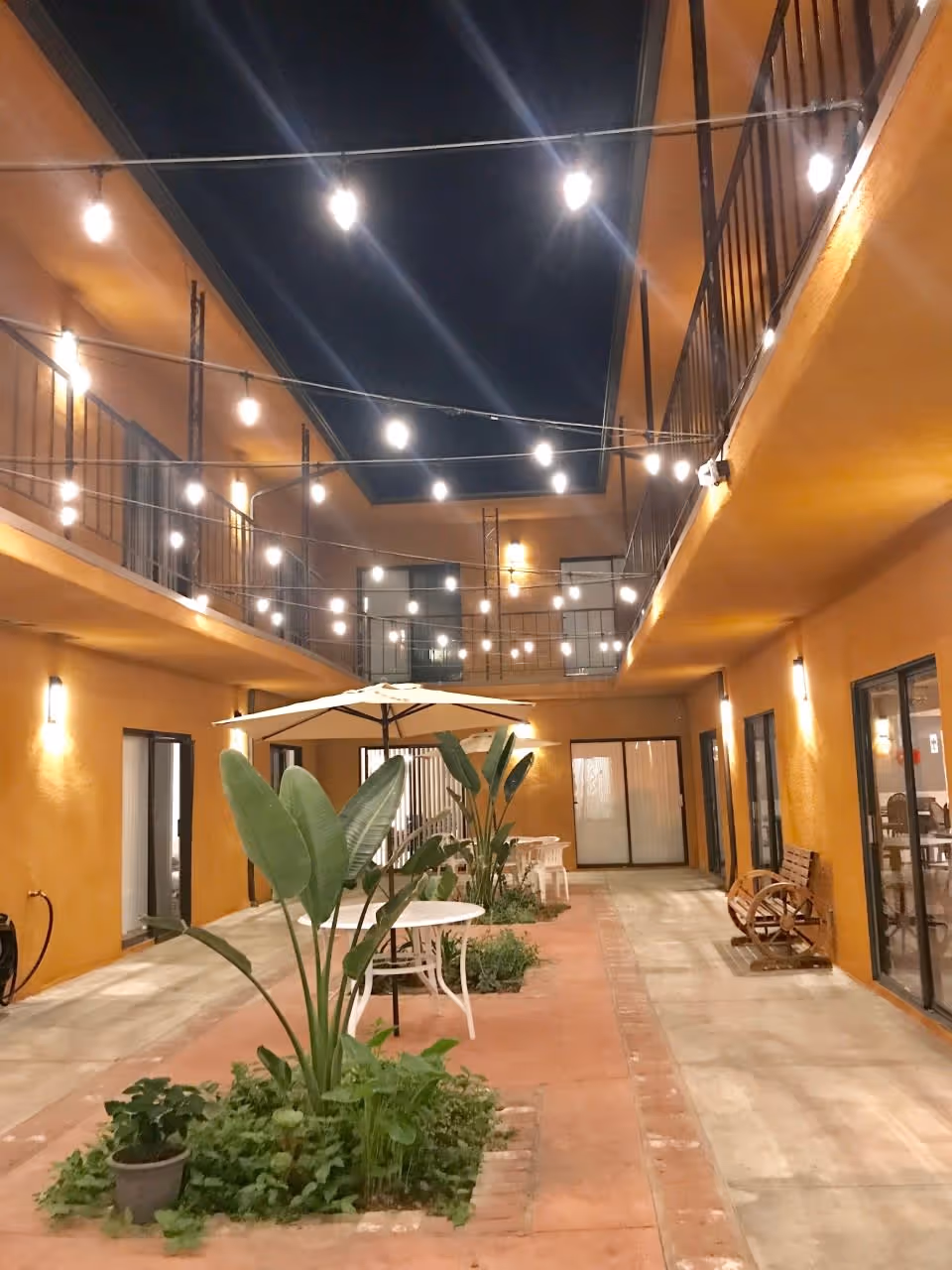 Two-story interior courtyard at night with hanging string lights, patio tables, umbrellas and plants between balconies.