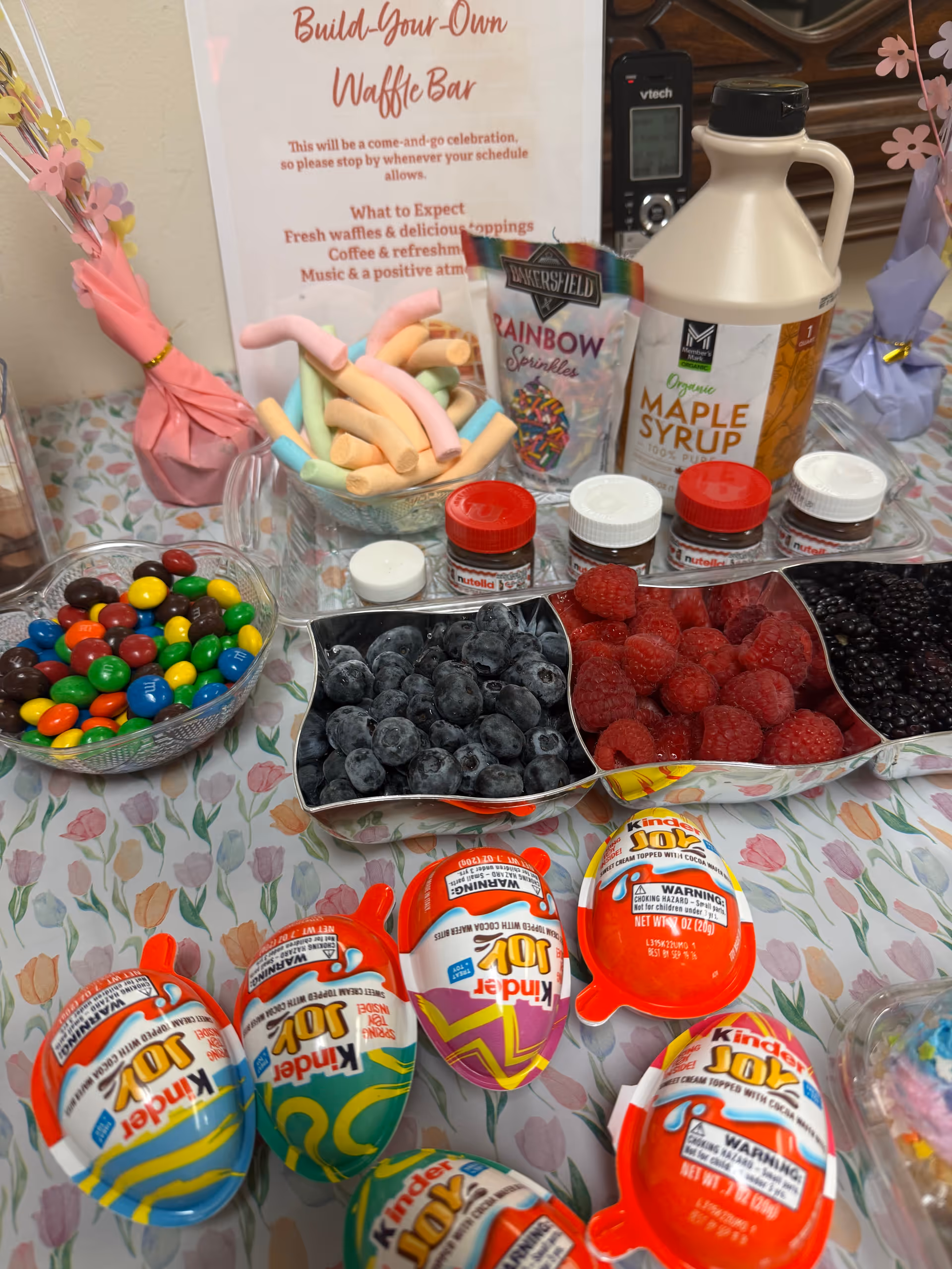 Build-your-own waffle bar with toppings, syrups, berries, and candy on a floral table.