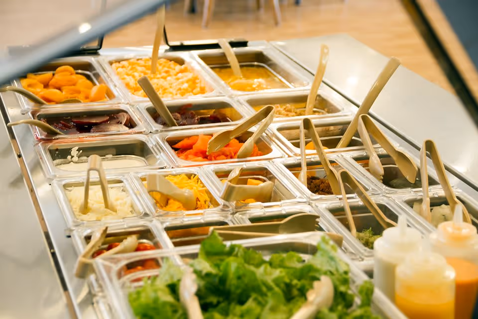 A salad bar with various fresh ingredients including lettuce, shredded cheese, diced onions, sliced carrots, beets, and other salad toppings in clear containers with serving utensils. There are also bottles of salad dressings on the side.