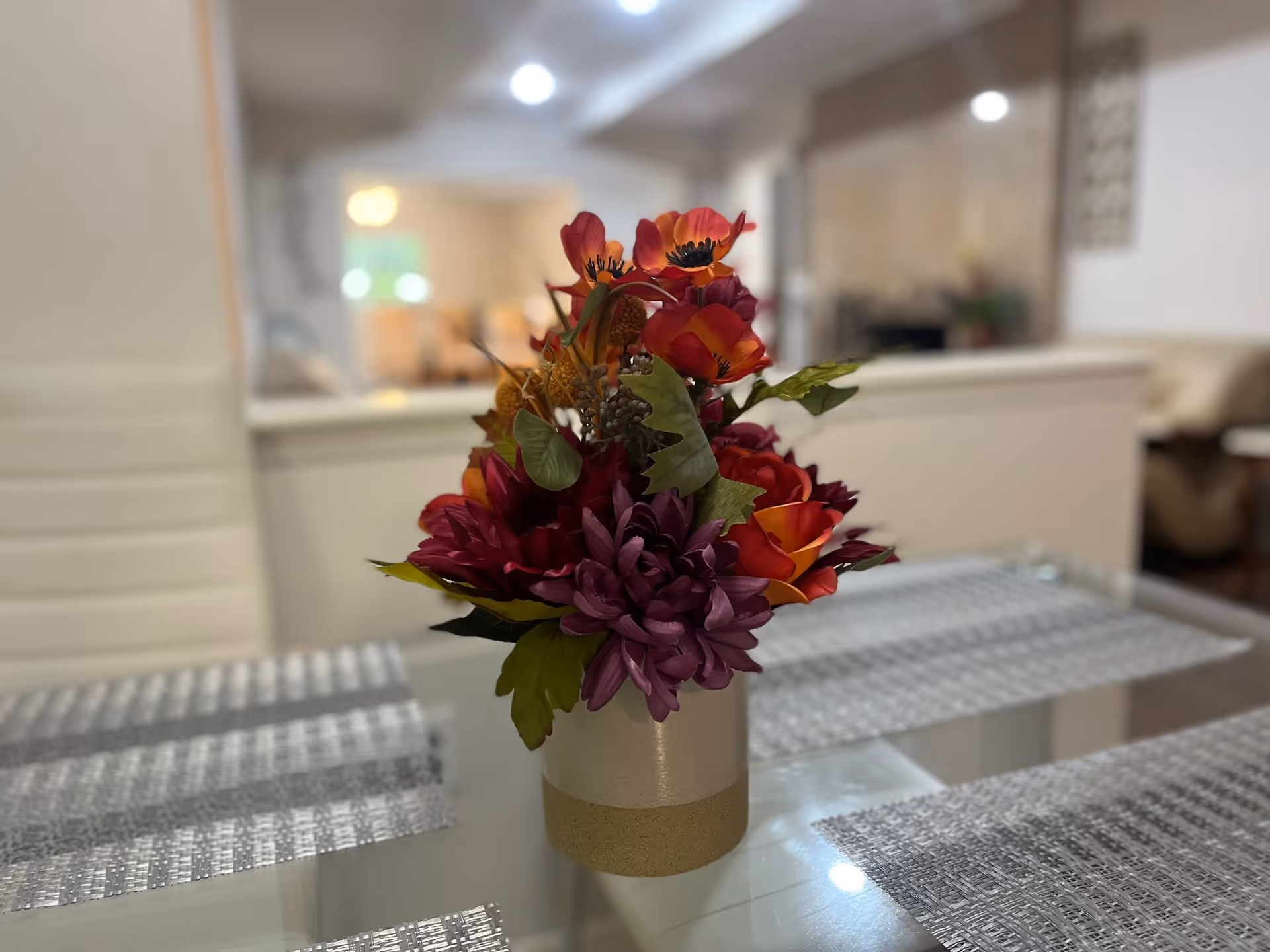A vase of colorful artificial flowers on a glass dining table with placemats and a blurred interior in the background.