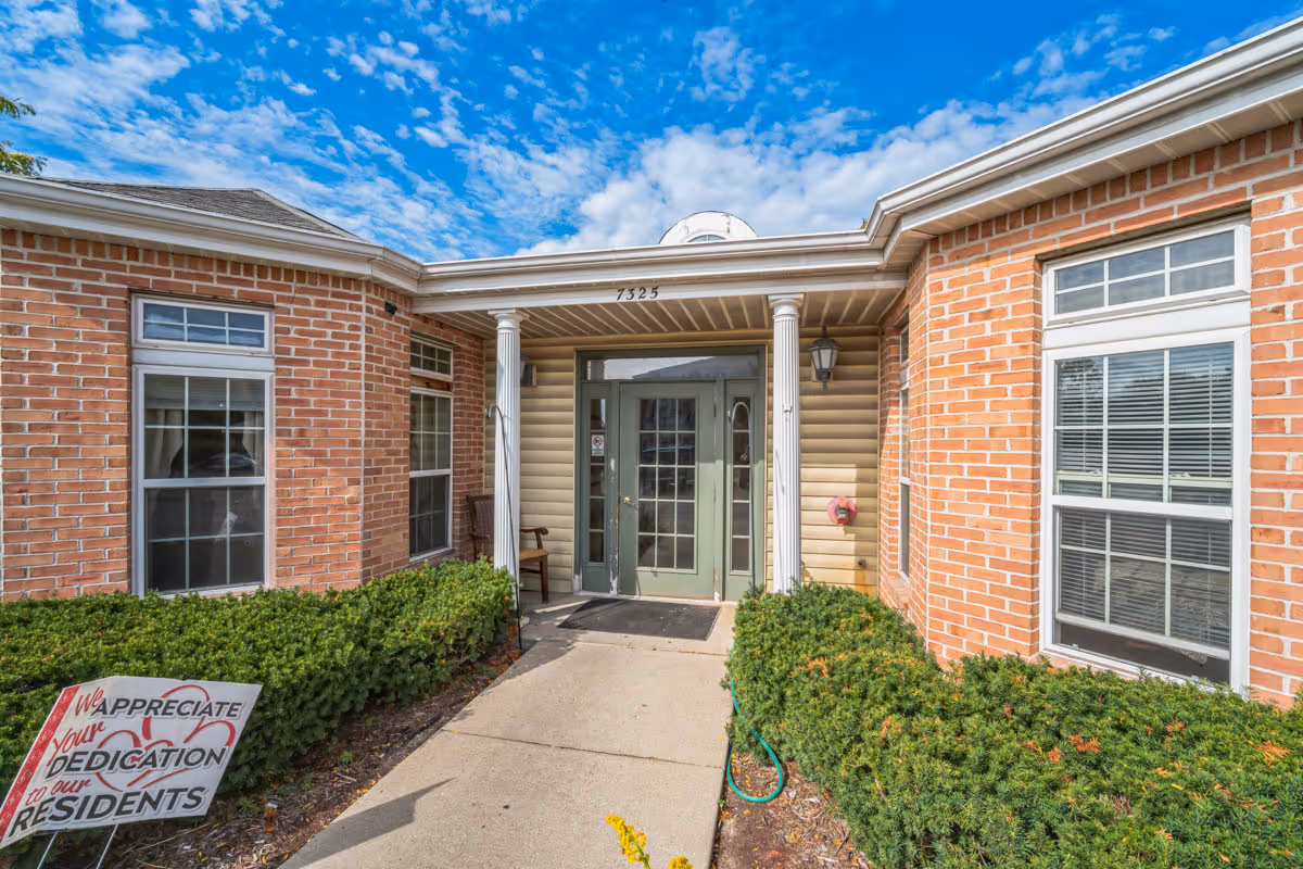 Entrance of a brick building with two white columns supporting a small roof over the doorway. The door is green with glass panels. There are windows on either side of the entrance and green bushes lining the walkway. A sign in the bushes reads 'We appreciate your dedication to our residents.' The sky is partly cloudy.