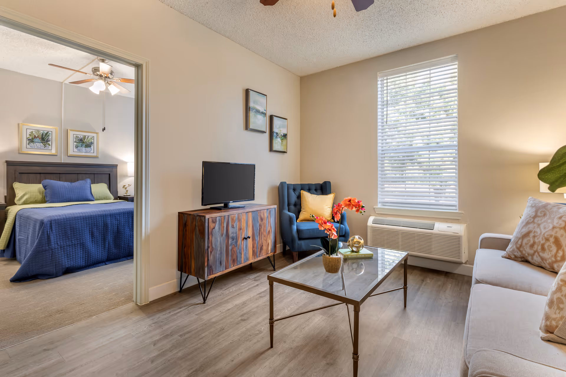 A cozy living room with a beige sofa adorned with patterned pillows, a glass coffee table with a potted orchid and decorative items, a blue armchair with a yellow pillow, and a wooden TV stand with a flat-screen TV. The room has light-colored walls and flooring, with a window covered by white blinds. Through an open doorway, a bedroom is visible featuring a bed with a blue quilt and green pillows, two framed botanical prints above the headboard, and a ceiling fan with lights.