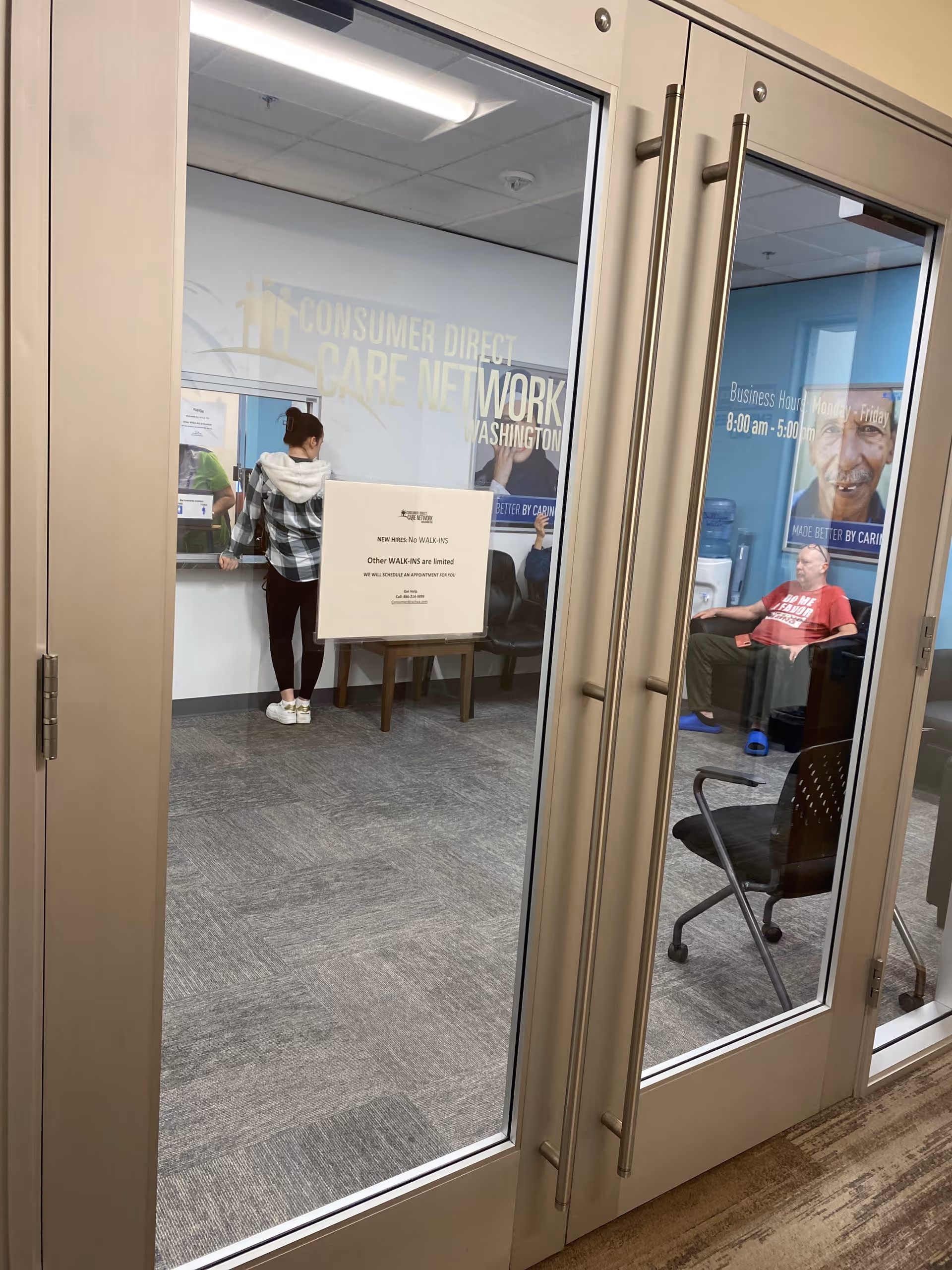 Glass doors open to a reception area with people at a counter and seating.