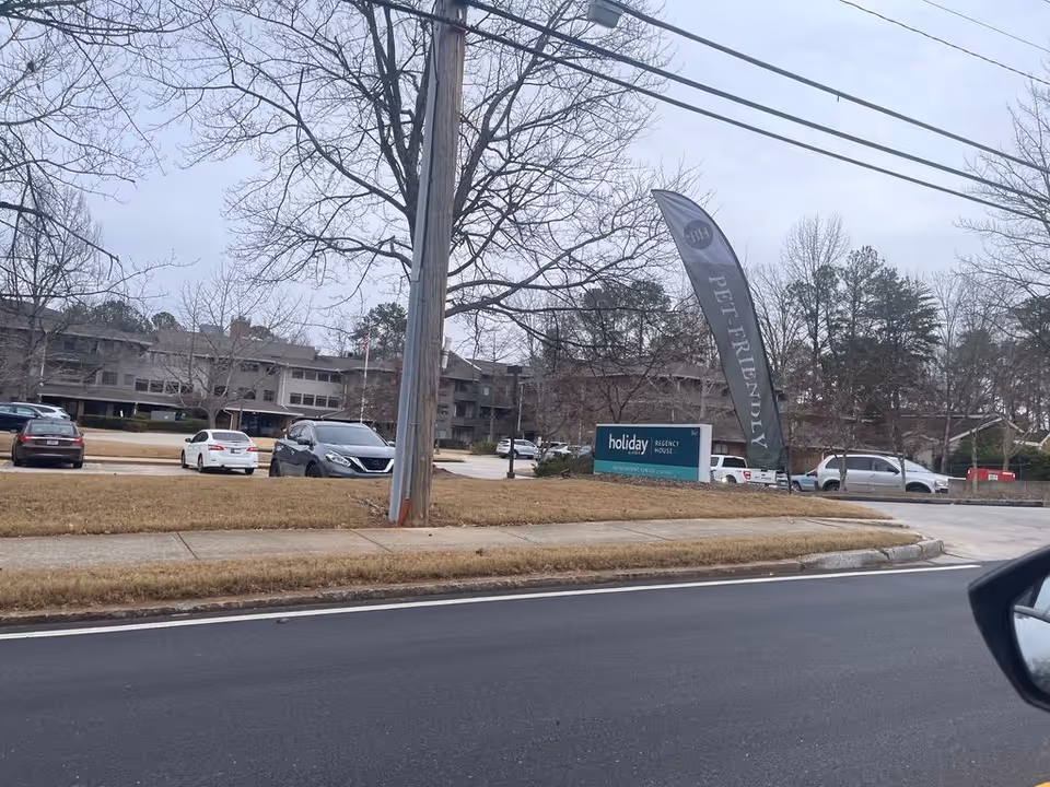 Street-front exterior of the Regency House senior living building with a parking lot, a 'holiday' entrance sign and a tall 'Pet-Friendly' flag.