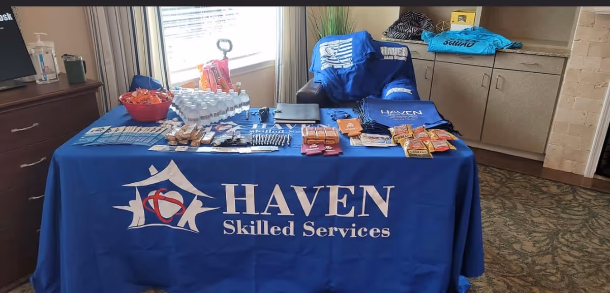 Promotional table with Haven Skilled Services branding, brochures, snacks, water bottles, and T-shirts indoors.