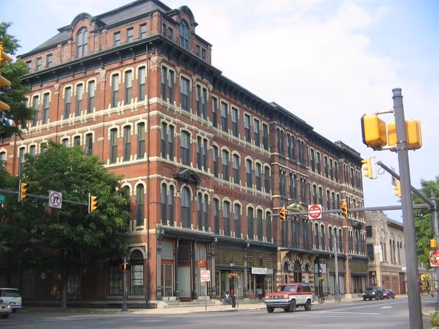 A large brick building with storefronts, traffic lights, and parked cars along a city street.