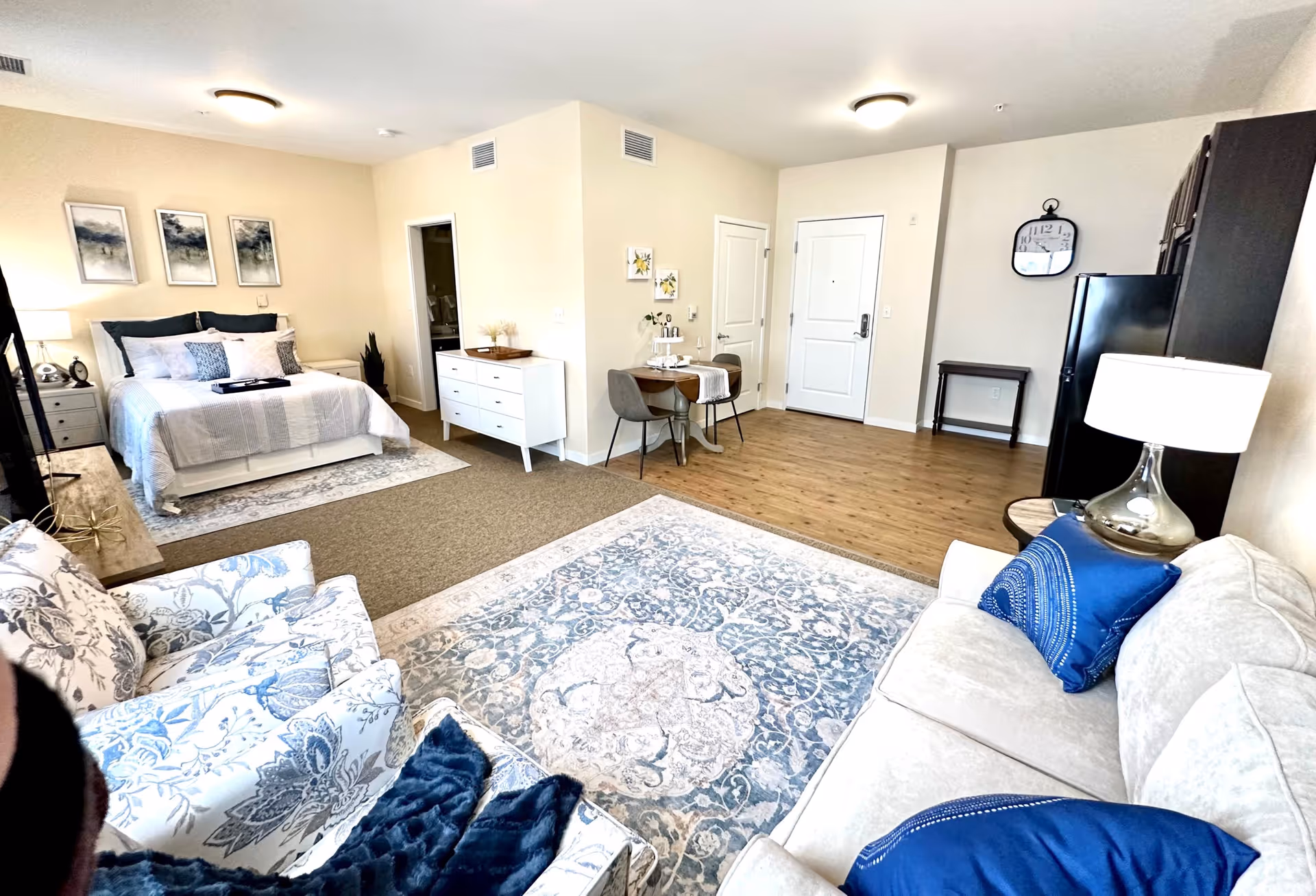 Open-concept senior living suite with bed, seating, dining area, and kitchenette.