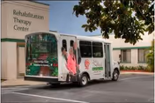 A white shuttle bus parked outside a rehabilitation center with signage and trees nearby.