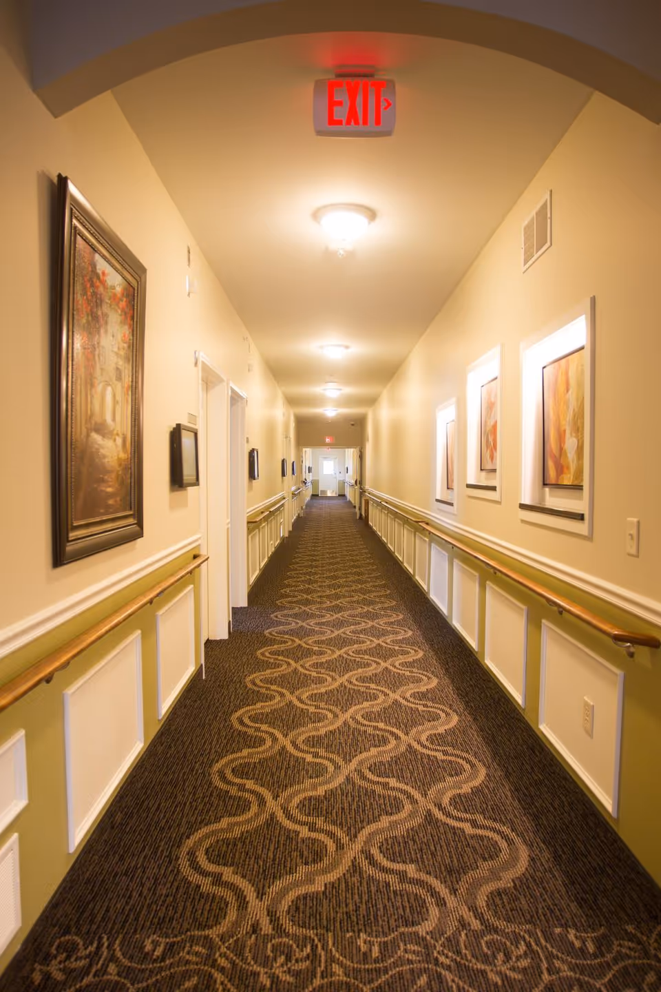 Long interior hallway in a senior living facility, lined with handrails, art, and ceiling lights.