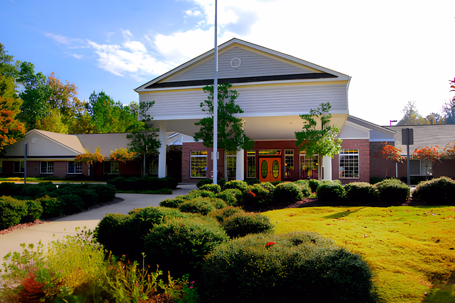 Front exterior view of a senior living facility named Serenity Empire with a covered entrance, surrounded by well-maintained bushes, trees, and a green lawn under a partly cloudy sky.