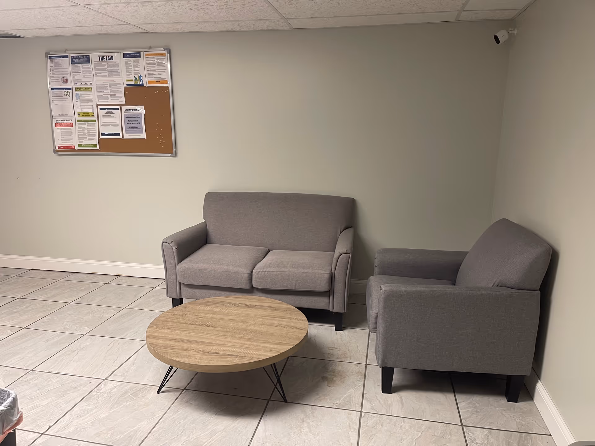 A simple waiting area with two gray couches, a round wooden table, and bulletin board.