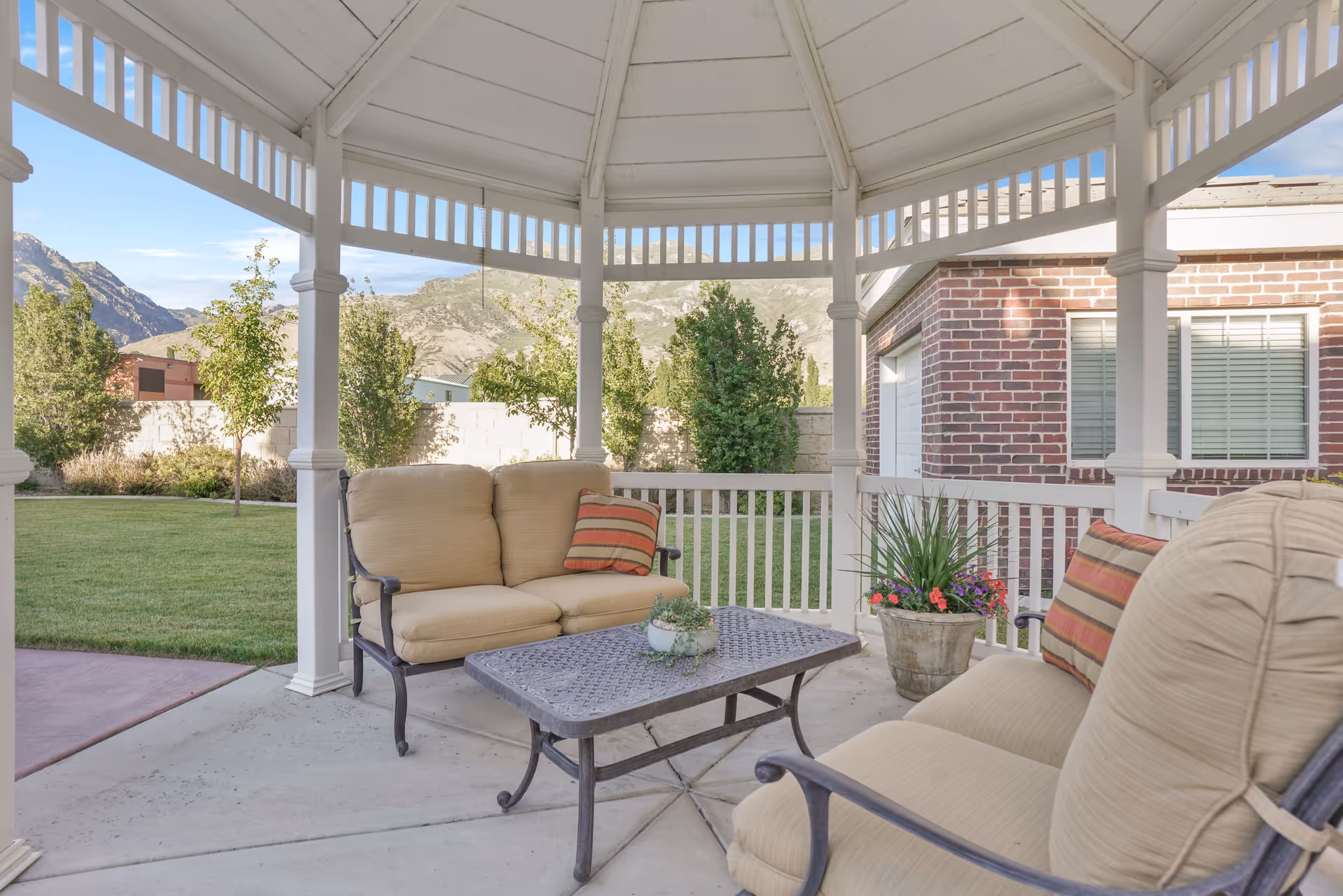White gazebo-style covered outdoor patio with beige cushioned seating, table, and potted plants.