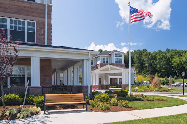 Front entrance of a senior living building with a covered drive, American flag, bench, and landscaped walkways.