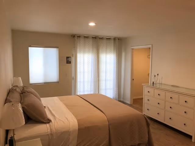 Bright bedroom with a neatly made bed with beige bedding, a white dresser, bedside lamps, and a curtained sliding door.