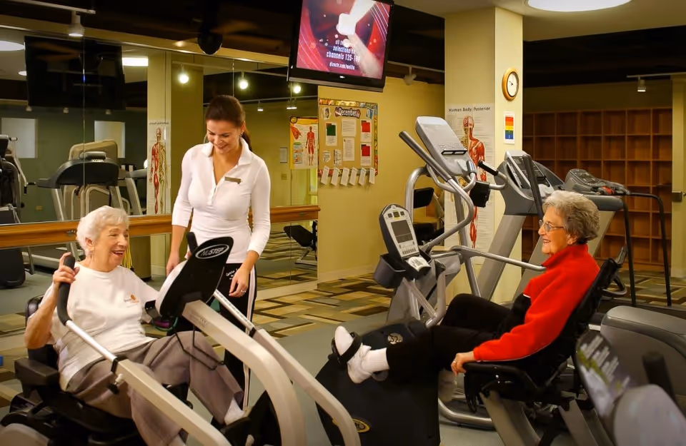 Two elderly women using recumbent exercise machines in a fitness room while a staff member assists them.