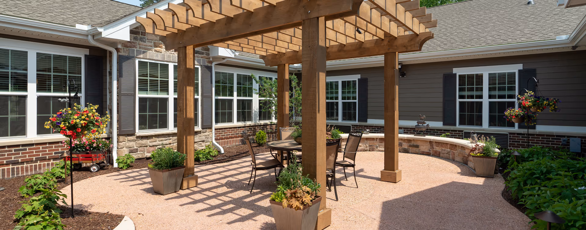 Outdoor patio area at Bickford of Chesapeake featuring a wooden pergola with a round table and four chairs underneath. The patio is surrounded by flower beds with various plants and hanging flower baskets, adjacent to the building with multiple windows.