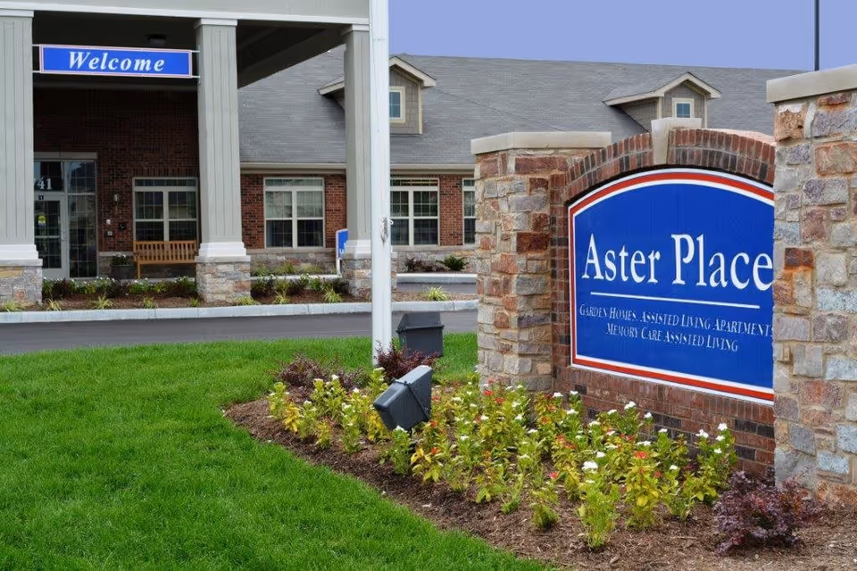 Entrance sign reading "Aster Place" in front of a brick senior living building with landscaping and a 'Welcome' sign over the driveway.
