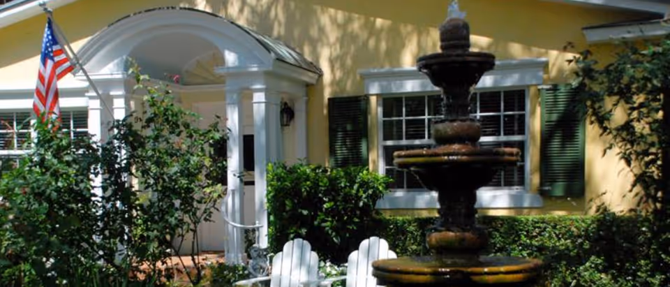 Front view of a yellow retirement residence with arched entry, fountain, and American flag.