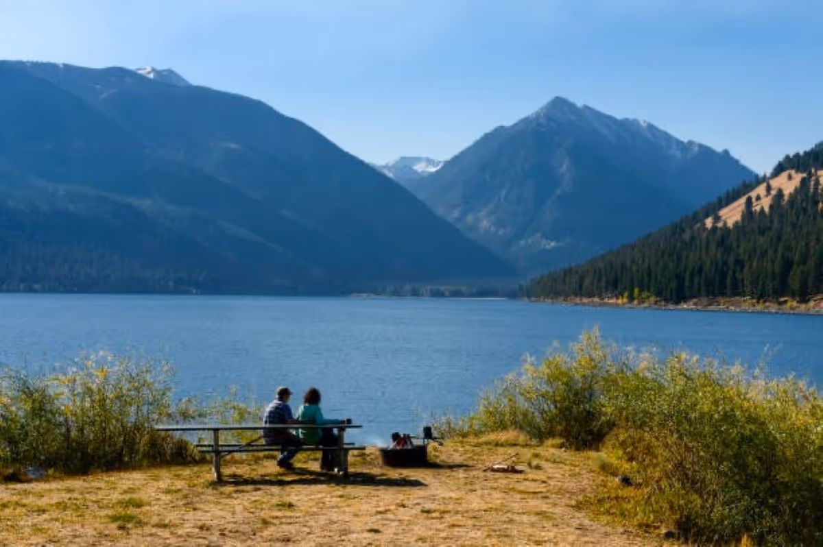 Two people sit at a picnic bench by a lake with mountains in the background.