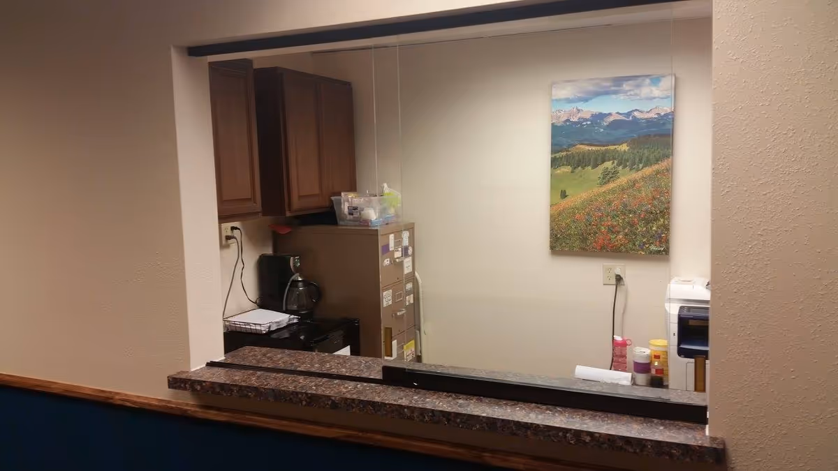 Interior view through a service window into a small kitchenette/office with cabinets, a coffee maker, a file cabinet, and a landscape painting on the wall.