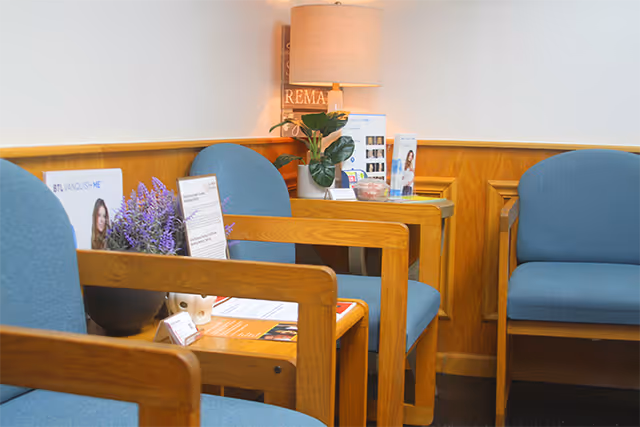 A small waiting area with blue upholstered chairs, wooden tables, a lamp, plant, and brochures.