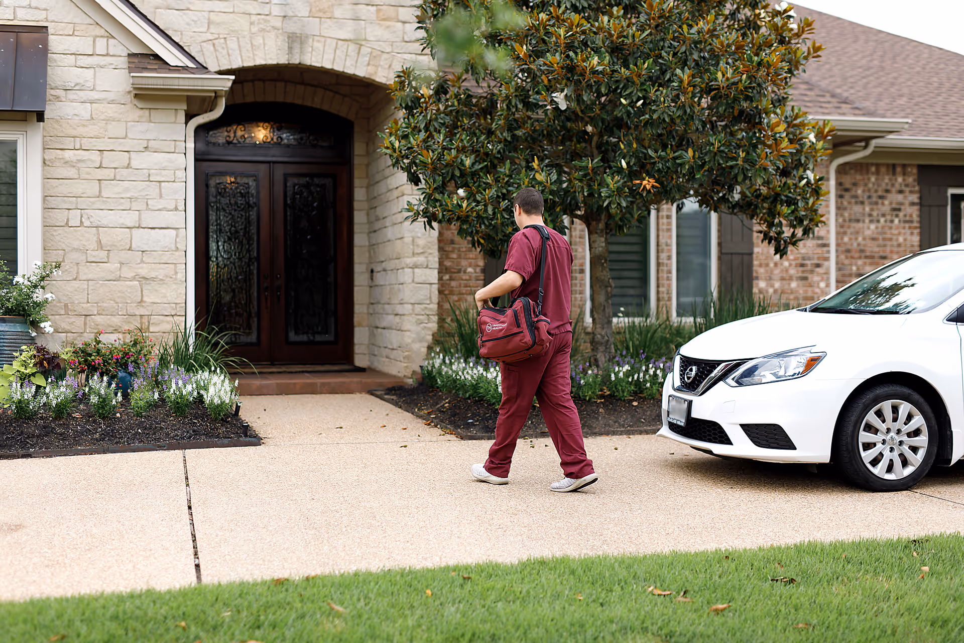 Healthcare worker in burgundy scrubs walks toward a door of a stone house, bag in hand